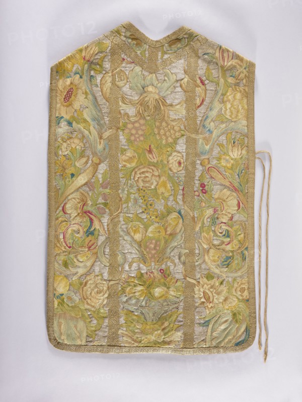 Chasuble, provenance unknown. Embroidered chasuble with birds, flowers, and fruit ornament; liturgical vestment used as a church parament. On a silver background, a multicolored pattern composed of a fruit vase, floral scrolls, grapes, and birds. Predominant colors: silver background and green, blue, and yellow ornament. Edges trimmed with gold braid with a geometric pattern. Internal divisions marked with strips embroidered with gold thread, imitating haberdashery. Cotton lining. Embroidery on linen canvas with multicolored silk and metal threads in two types: silver silver plate woven on a white silk base and gold gold plate woven on a yellow silk base. Metal thread embroidery in a laid flat stitch; colored embroidery in split, flat, and knot stitches. 18th century, Poland. Length 105 cm, width 64 cm back., An ornate textile panel with a pointed top and rounded bottom displayed against a light background, featuring an overall palette of gold, pale yellow, soft green, muted blue, crea...