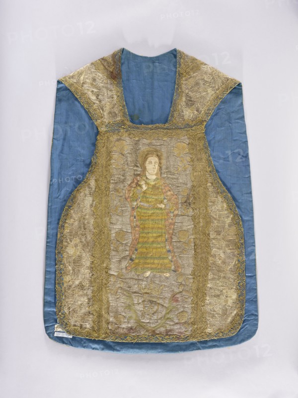 Chasuble, Madonna and Child, St. Bridget 1303–1373, Slepowron coat of arms, coats of arms, religious scenes, saints, floral ornaments, flowers, liturgical vestments, plant ornaments, church paraments, sacred art, churches and chapels furnishings. Chasuble made of solid fabric with a floral pattern, with embroidered columns. On the front column is the figure of St. John the Evangelist holding a chalice with a snake; below is a cartouche with the Slepowron coat of arms, around the cartouche the letters TLSD. On the back column is Mary and Child on a crescent; below is a kneeling Saint Bridget with an inscription around her head: SM BRI/GITTA. Stylized floral motifs embroidered in the background of the columns. The columns and sides of the chasuble are trimmed with gold bobbin lace. 17th century, Spain fabric?, Poland embroidery?. Liturgical vestment. Length 100 cm, width 69 cm back., A garment laid flat against a pale background, predominantly blue with a sheen to the fabric, featuring a...
