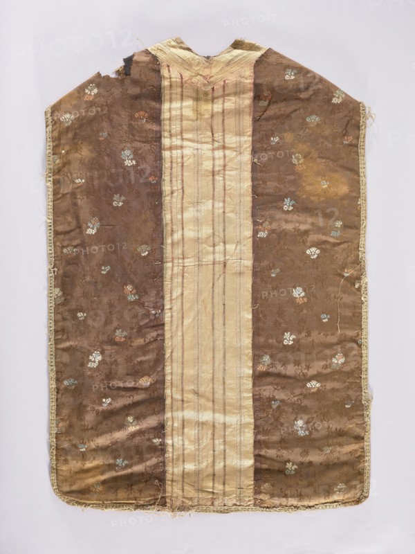 Chasuble, liturgical vestment; 18th century. Silk columns with a pattern of pink and silver stripes. Sides made of brown fabric with floral motifs, partly in damask technique and partly brocaded with colorful silks: sky blue, pink, and cream. Columns from France, sides from Venice. Length 112 cm, width 70 cm back., A rectangular garment laid flat with a central vertical panel in a pale beige color bordered by a wider brown fabric on each side, the brown areas patterned with small floral motifs in muted colors including light blue, pale green, white, and rusty orange; the central panel has faint vertical stripes in slightly darker beige and reddish tones; the garment shows frayed edges and worn areas, with a scalloped neckline at the top and a narrow decorative trim in a light beige running along the outer sides and bottom; the overall colors visible are brown, beige, light blue, pale green, white, and rusty orange.