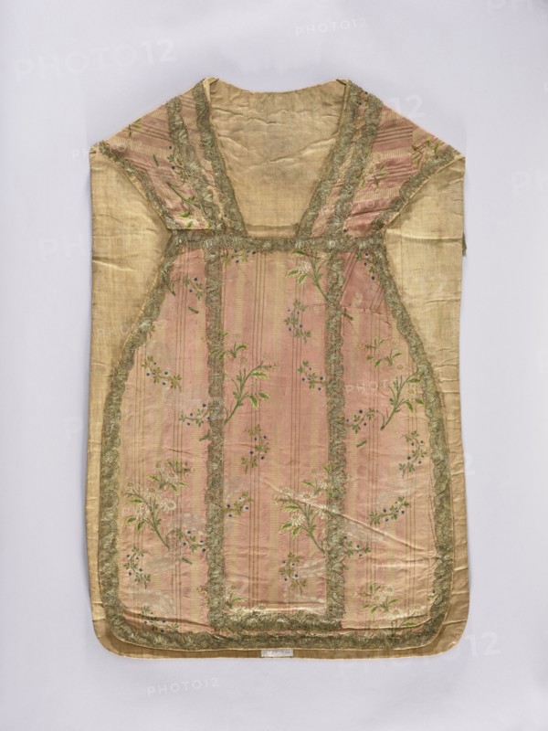 Chasuble, liturgical vestment;, noted. A large floral pattern and lace motif are set against a raspberry background. The dominant colors in the ornament are brown, silver, and green. The entire piece is trimmed with period lace. Divided into columns with sewn-on gold lace. 2nd half of the 18th century, Lyon, France. Liturgical vestment. Length 99 cm, width 66 cm back., A sleeveless garment laid flat with a rectangular body and a V-shaped neckline, made of a pale gold fabric with a central panel in a muted pink tone that features vertical stripe textures and a floral pattern; the pink panel is bordered by a scalloped trim in a metallic gold color, and the floral motifs include green leaves and small blue accents; the shoulder and side sections are pale gold matching the outer fabric, and the overall piece shows gentle creases and soft folds against a light neutral background.