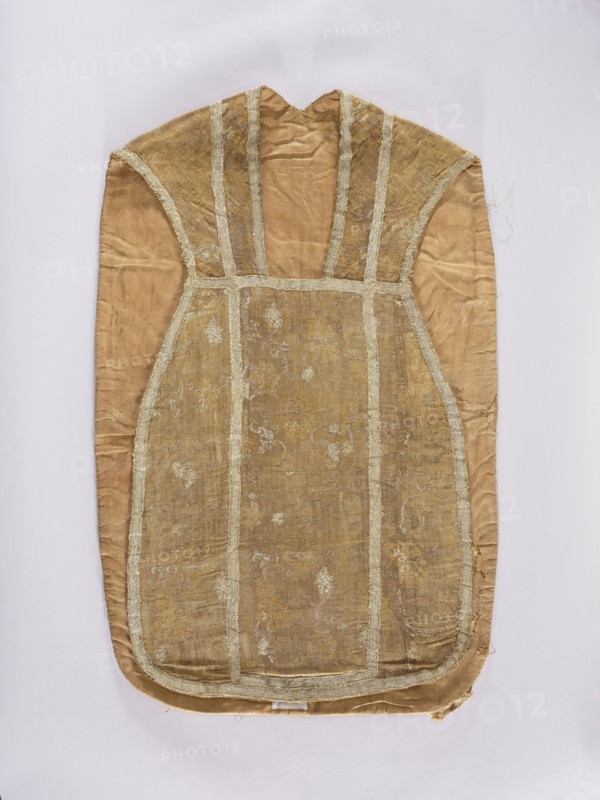 Chasuble, liturgical vestment, 18th century, France. Division into columns with sewn-on silver braid; edges trimmed with the same braid. Features flowers and plant ornaments; used as church paraments/sacred art for churches and chapels. Length 101 cm, width 63 cm back., An elongated fabric garment laid flat, with a rounded lower edge and a narrow upper neck opening, showing a central panel bordered by vertical and horizontal bands; the fabric appears worn and has a textured brocade pattern visible in places. The visible colors include shades of beige, tan, gold, and light cream, with the central panel slightly darker and more golden than the surrounding tan fabric. The bordering bands are a lighter cream color with frayed edges, and there are areas of subtle sheen and irregular wrinkles across the material. The garment edges show some wear and loose threads, and the surface displays small patched or faded spots where the pattern is less distinct.