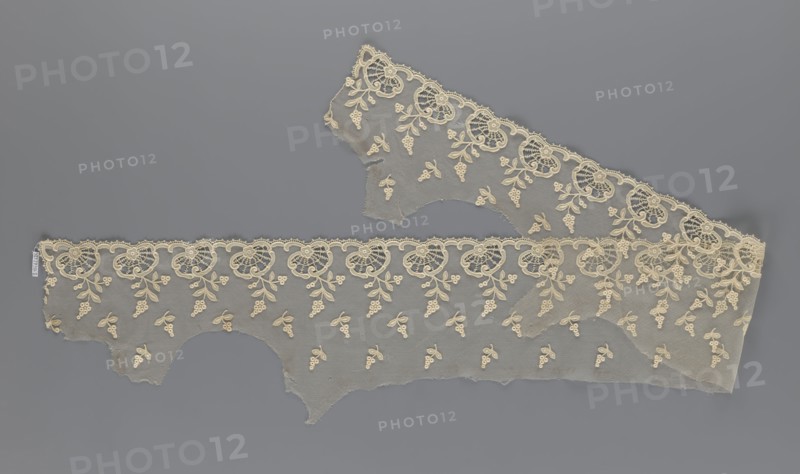 Fragment of lace, lace, plant ornaments, slightly serrated lower edge with shell motifs and sprouting twigs, above it projections of smaller twigs, 2nd half of the 19th century, Europe, lace, width 20 cm, length 161 cm, A length of delicate lace fabric laid flat on a gray background, showing a scalloped decorative edge and floral motifs worked into a fine net ground; the lace appears in shades of cream and off-white against the medium gray backdrop, with a repeating pattern of circular floral rosettes along the scalloped border and small clustered floral sprays scattered across the mesh, and with irregular torn edges and a few overlaps where the lace layers cross.