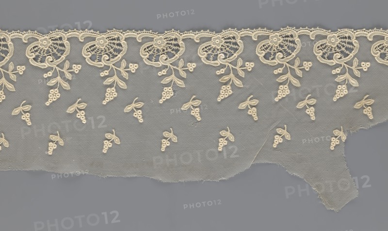Fragment of lace, lace with plant ornaments, slightly serrated lower edge with shell motifs with sprouting twigs and above them projections of smaller twigs, 2nd half of the 19th century, Europe, width 20 cm, length 161 cm, A length of delicate lace in cream tones lies against a flat gray background; the lace features a scalloped upper edge with repeating ornate floral motifs and a mesh net ground sprinkled with a few small clustered berry-and-leaf designs, and the lower edge is uneven and slightly frayed.
