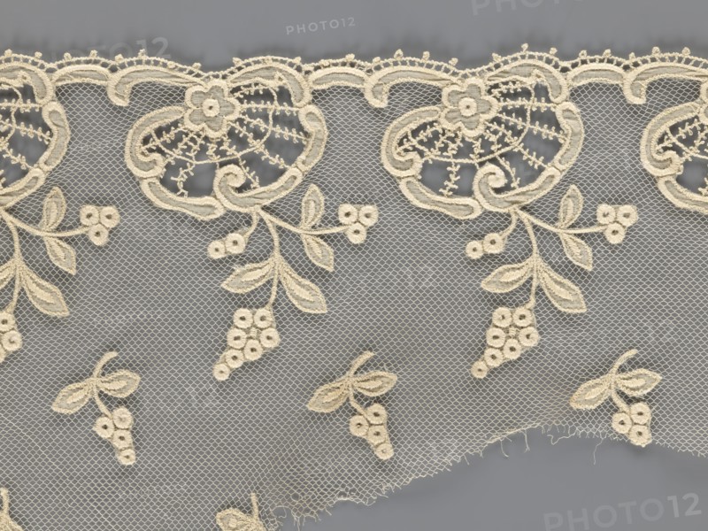 Fragment of lace, lace with plant ornaments, slightly serrated lower edge with shell motifs and sprouting twigs, above it projections of smaller twigs; 2nd half of the 19th century, Europe; lace, width 20 cm, length 161 cm, The image shows a piece of lace fabric with an intricate floral and vine pattern stitched in a cream color against a gray background. The lace features a scalloped top edge with embroidered flower motifs and looping ornamental shapes, and a net-like mesh fills the background. Several hanging clusters of embroidered berries and leaves extend downward, and a few small detached floral clusters are scattered across the lower portion. The lace has delicate openwork details and a slightly frayed lower edge.