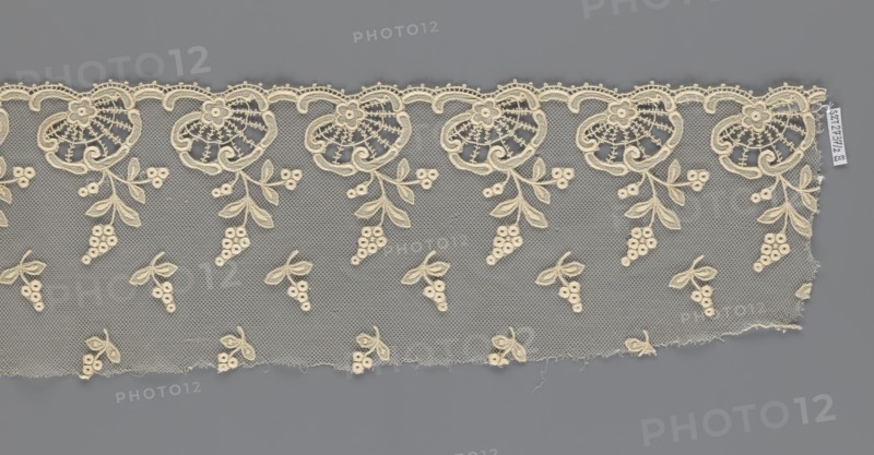 Fragment of lace, creator unknown, lace of, plant ornaments, slightly serrated lower edge with shell motifs and sprouting twigs, above it projections of smaller twigs; 2nd half of the 19th century, Europe; lace, width 17 cm, length 86 cm., A strip of delicate lace with an open net background in gray and cream tones, featuring a repeating floral border along the top with ornate fan-shaped blossoms and scalloped edging in cream, and a pattern of small clustered berry motifs and paired leaves scattered across the lower area; the lace shows frayed edges and loose threads along the bottom and right sides, and the overall colors visible are cream and gray.