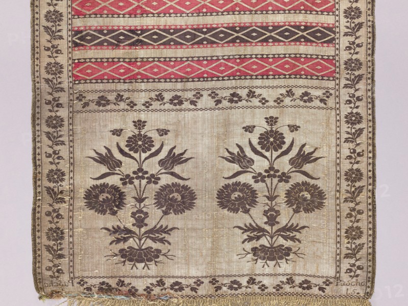 Kontusz sash, Paschalis Jakubowicz Manufactory Warsaw; belt manufacturer; 1788-1794. Solid kontusz sash, double-sided. Bipartite heads with a flower bush pattern. Alternating transverse stripes, plain and with geometric ornamentation. Edge with a floral tendril motif. Plant ornaments, geometric ornaments, flower bushes, floral ornaments. Colors: gold, silver, blue, yellow-green, light red, brown, light blue. Sew-on fringe, silver, with gaps. Ornament: - Heads: two motifs of rooted flower bushes. - Center: alternating transverse shelves, smooth and with rhombus motifs. - Borders: vertical and horizontal with different floral tendril motifs; checkerboard and rhombus. Colors placement: - Front: background of heads, borders and smooth shelves silver, ornament brown; background of the shelves with rhombuses alternating red and brown, silver ornament. - Reverse right: only smooth shelves alternating red and brown. Signed in the heads: Pascha/Nov. Weave density: approx. 19/34 x2. Date: 1788–1...