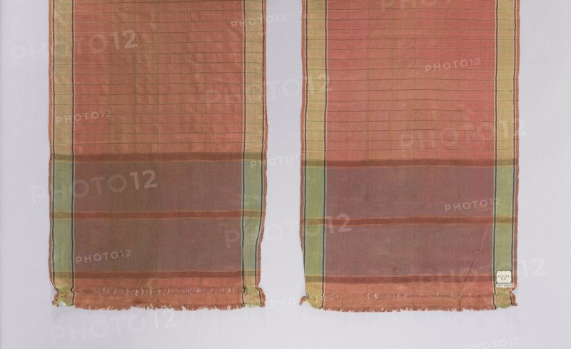 Kontusz belt, Polish/Ukraine Volhynia? origin, mid-18th century. Men's clothing accessory. Silk with smooth heads, half-cut by a 1 cm wide line; smooth stripes separated by narrow stripes; along both sides a green trail 3 cm wide. Length 240 cm, width 42.5 cm., A pair of long rectangular fabric panels hangs side by side against a pale background, each panel showing a horizontal striped pattern with a dominant warm reddish-orange field transitioning to a muted brownish band near the lower portion; both panels have vertical border stripes in pale yellow and green with thin dark lines, and the fabric appears semi-sheer with a subtle grid texture. The lower edges of the panels show frayed hems with loose threads and small gathered corners, and there is a small pale patch near one lower edge. Visible colors include reddish-orange, brown, pale yellow, green, dark thin accent lines, and the pale neutral background.