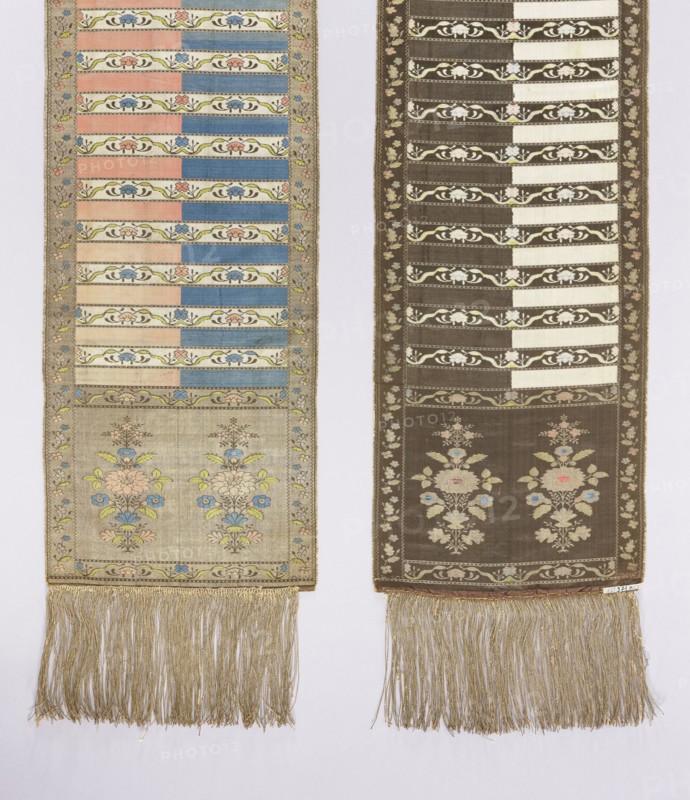 Kontusz sash. Semi-solid, four-sided sash with plant and flower-bush ornament; bipartite heads with a motif of a stylised flower tree growing from a miniature mound. One side: white and brown background with brown heads; other side: blue, crimson and gold heads. Pattern on the gold heads is multicolored; on the brown heads is off-white. Signed at the bottom of the heads, lying down, FS. 18th century, Lyon. Polish sash used with national costume; men's fashion accessory. Length 294 cm, width 30 cm., Two long vertical woven panels hang side by side against a pale background. The left panel has a light beige ground with horizontal bands of alternating pastel pink and blue, narrow decorative floral bands in green and cream, a lower section featuring stylized bouquets in pale pink, blue, yellow, and green, and a fringed bottom of golden threads. The right panel has a darker brown background with similar horizontal striped sections in cream and brown, decorative floral bands in cream and gre...