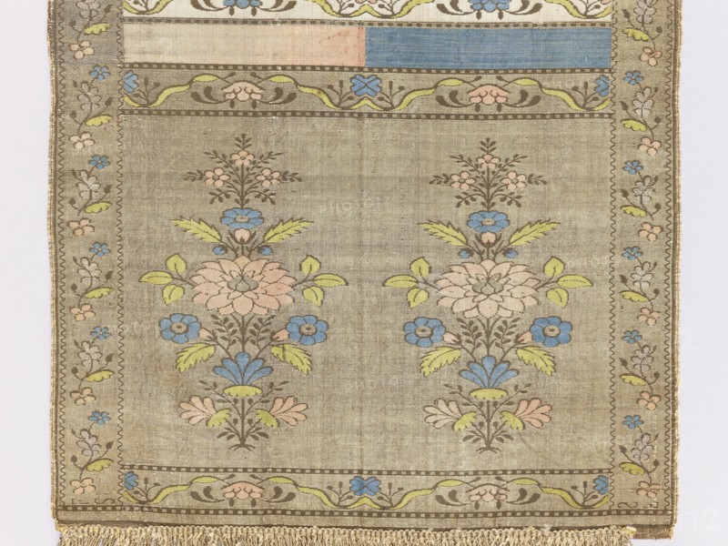 Kontusz sash, semi-solid sash with flower bushes ornament, four-sided sash with plant ornaments, Polish sash used in national costume, men's clothing accessory. Bipartite heads with a motif of a stylised flower tree growing from a miniature mound: on one side a white and brown background with brown heads, on the other blue and crimson and gold heads. The pattern on the gold heads is multicolored, and on the brown heads is off-white. Signed at the bottom of the heads, lying down, FS. 18th century, Lyon. Length 294 cm, width 30 cm., A rectangular woven textile with a muted beige background featuring a repeating floral motif in pastel colors; a pair of vertical floral sprays with stylized blossoms in pale pink and blue, leaves in light green, and outlining in brown sit side by side within a bordered field, accompanied by smaller floral clusters above; a decorative inner border with flowing vine shapes and small flowers in blue, pink, and green surrounds the central area, and a narrow oute...