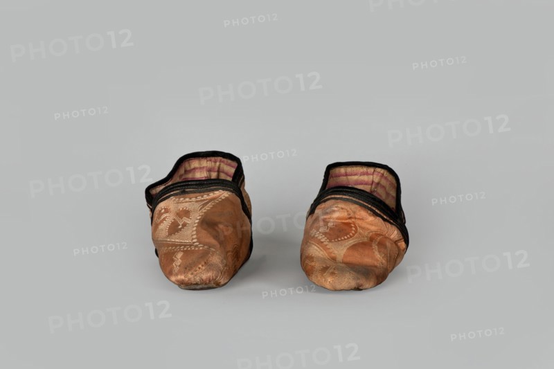 Slippers, origin China, 19th century. Women's footwear: slippers with haberdashery decorations. Lining of the upper and sole made of pink striped canvas; illegible rectangular stamp on the lining. Machine sewing supplemented by hand. Leather sole with a semicircular under-heel piece, tapering sharply to a pointed toe; second layer of leather under the heel nailed with metal nails. Height 5.3 cm, length 17 cm., A pair of soft slippers placed side by side facing forward on a neutral light gray background, each slipper showing a worn orange-brown fabric exterior with decorative stitched patterns, black trim around the opening, and an inner lining with faded pink and beige stripes; the slippers appear slightly misshapen and creased with visible areas of darker discoloration on the toes, and the overall color palette includes orange-brown, black, pink, beige, and gray.