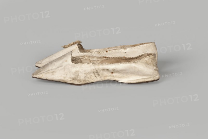 Women’s slippers. Maker: Johann Dyck, shoemaker’s workshop in Vilnius ca. 1800–ca. 1850. Flat women’s slippers made of white full-grain leather with flat-cut toes, decorated with a rosette of white grosgrain ribbon one rosette preserved. Symmetrical leather soles, white canvas lining and endpaper. A printed company label is glued onto the endpaper of one slipper: “Johann / Dyck / …auf der / Schloss-strase / nr … in / VILNIUS.” Date: 1st half of the 19th century 19th century. Type/place of origin: Vilnius. Category: footwear, clothing; women’s footwear/slippers. Dimensions: height 6 cm; length 24 cm., A pair of worn slip-on shoes made of a light material, shown from the side and overlapping, with pointed toes and low flat soles; the shoes display visible creases, scuffs, and frayed edges, especially around the openings, and the interior lining is partly exposed; the dominant colors are off-white and beige with areas of darker dirt and brownish discoloration, and the soles appear darker ...