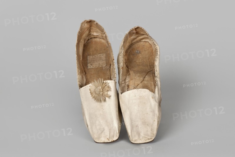 Women’s slippers, maker: Johann Dyck Vilnius; shoemaker’s workshop; ca. 1800–ca. 1850. Flat women’s slippers made of white full-grain leather, 1st half of the 19th century. Flat-cut toes, decorated with a rosette of white grosgrain ribbon one rosette preserved. Symmetrical leather soles. White canvas lining and endpaper. A printed company label is glued onto the endpaper of one slipper: “Johann / Dyck / …auf der / Schloss-strase / nr … in / VILNIUS.” Height 6 cm; length 24 cm. Type/place of origin: Vilnius. Materials/subjects: haberdashery decorations, company labels, leather goods, slippers, footwear, women’s footwear, women’s slippers, fashion, clothing accessories., A pair of worn slip-on shoes shown from above on a plain background, the shoes are primarily off-white with visible areas of beige and brown on the interiors, the outer material appears smooth with some scuffs and slight discoloration, one shoe has a decorative rosette made of the same off-white material near the opening...