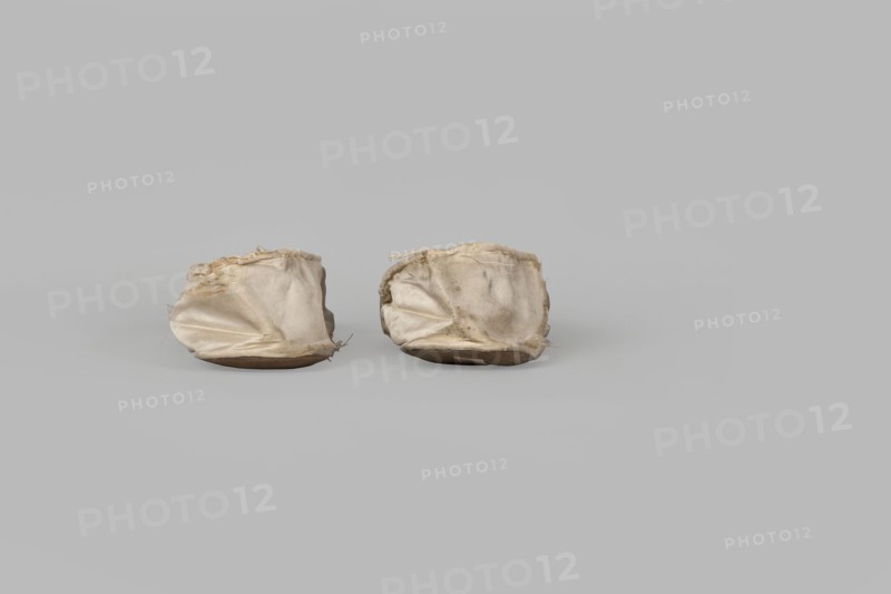 Flat women's slippers of white satin, top edge finished with ribbon. Flat-cut toes, symmetrical leather soles. Endpaper and back lining of white leather, front lining of canvas. On the endpaper of both slippers there is an octagonal, oblong company label: Jan Kólazenski / in Kalisz / at Market Square No. 18. Maker: Jan Kólazenski, Kalisz; shoemaker's workshop; ca. 1830–1850. Women's footwear, clothing accessory; height 6 cm, length 23.7 cm., A pair of small fabric items sit side by side on a flat surface, each appearing soft and slightly collapsed with wrinkled, worn cloth that has frayed edges; the fabric shows shades of off-white, beige, and light tan with some darker smudges, and each item rests on a low, rounded base that is brown; the background and surface are a uniform light gray.