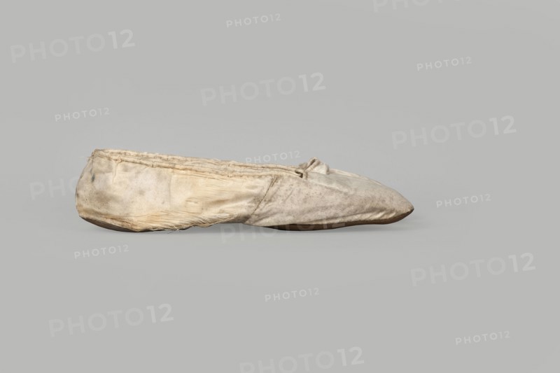 Flat women's slippers of white satin, top edge finished with ribbon. Flat-cut toes, symmetrical leather soles. Endpaper and back lining of white leather, front lining of canvas. On the endpaper of both slippers there is an octagonal, oblong company label: Jan Kólazenski / in Kalisz / at Market Square No. 18. Made by Jan Kólazenski, Kalisz; shoemaker's workshop, ca. 1830–1850. Women's footwear, slippers; height 6 cm, length 23.7 cm., A worn fabric slipper sits sideways on a plain background, showing a rounded toe and a low, flat sole; the fabric appears creased and frayed with visible stitching along the top edge and a small tied bow near the opening. The slipper's surface shows uneven discoloration and spots, with areas of dirt and wear, and some loose threads along the seams and lower edge. The visible colors are off-white, beige, light brown, and gray, with a darker brown sole.