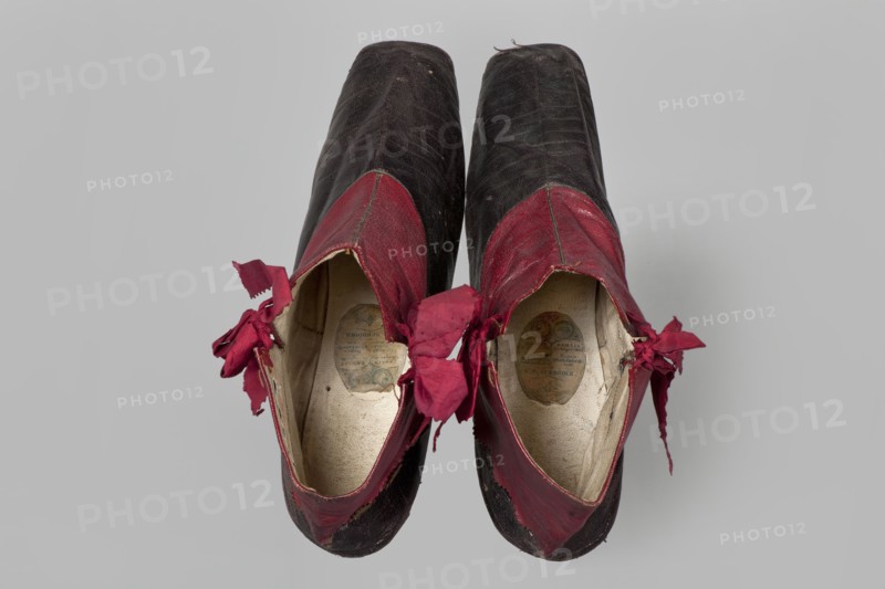 Women's ankle boots made of graphite full-grain leather with red leather uppers tied on the outside and inside with a red ribbon. Thin, symmetrical leather soles with bevelled toes and no heels. Canvas lining, white leather endpapers. Oval company label on the endpaper: Berlin / 23 Jäger str / Fabrique de chassures / pour les Dames / Berlin / 23 Jäger str / Damen / Schuh-fabrik von / CP Schröder. Upper part machine-sewn, lining hand-sewn. Circa 1850, Berlin. Height 10 cm, width 5.5 cm, length 25.5 cm. Women's ankle boots; fashion, footwear, women's footwear, fashion accessories., A pair of worn low-cut shoes photographed from above, showing the tops and insides; the shoes have dark upper bodies with prominent red sections around the openings and frayed red fabric bows tied at the sides; the interiors are a light beige color with visible worn insoles; the exterior shows scuffs and creases in dark and red areas, with small bits of loose threads; the background is a plain light gray.