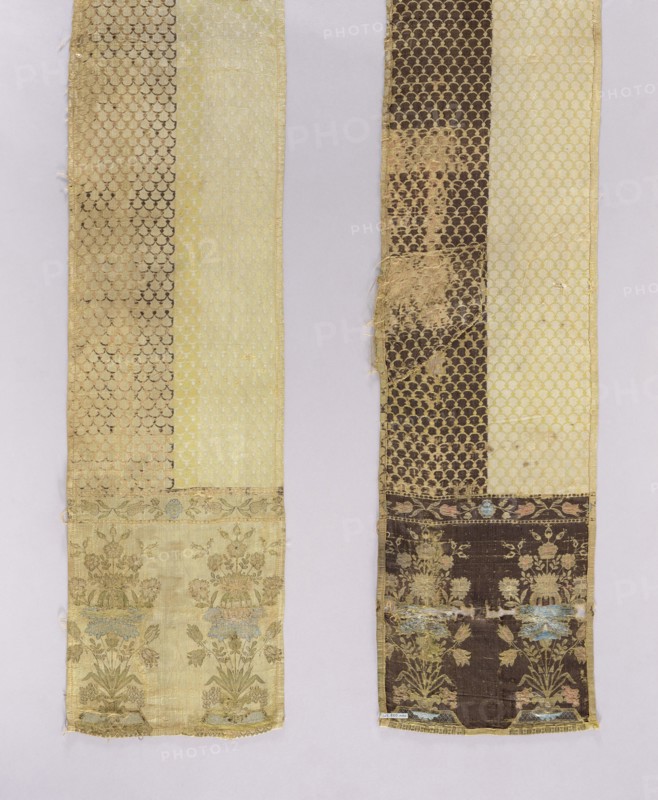 Kontusz belt, flower bouquets ornament, kontusz belt men's national costume accessory, Poland, carp scale ornament, bipartite heads with a floral bouquet pattern on a base, colors: ochre, blue, green, brown, yellow, dated 1768–1780, place of origin Kutkorz, clothing accessory, length 290 cm, width 29 cm, A close-up of two long vertical textile strips laid side by side on a pale background, each strip showing decorative woven patterns and areas of wear; the left strip features a pale creamy yellow panel and a darker golden-beige patterned panel with a repeating scallop motif, and a lower section showing a floral border with muted green and pale blue accents on a light yellow-beige ground; the right strip shows a dark brown patterned panel with a repeating scallop motif alongside a pale yellow panel, and a lower floral border with faded green and pale blue details on a dark brown ground, with some fraying and worn patches revealing lighter threads.