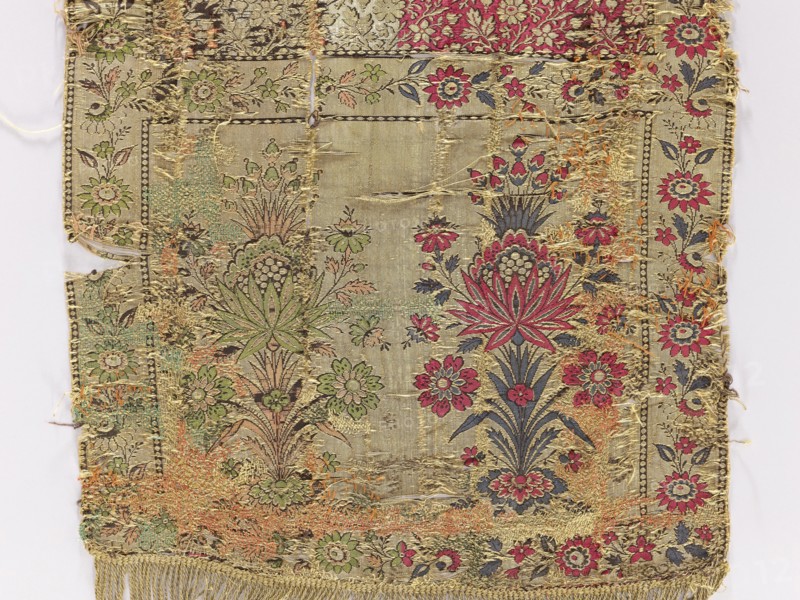 Kontusz belt, Polish clothing accessory. 2nd half of the 18th century, Lyon, France. Four-sided, bipartite heads with a floral bouquet pattern; continuous pattern of flower branches with tendrils of large flowers. Sewn-on gold fringe. Materials/colors: gold, light green, pink, navy blue, cherry red, black. Length 174 cm, width 36 cm., A worn rectangular textile fragment with a floral pattern, showing an ornate central panel flanked by decorative borders; the fabric background is a beige or light gold tone with visible areas of fraying and thread loss. The design includes stylized flowers and foliage in shades of red, pink, green, dark blue, and muted brown, with gold or yellow metallic threads woven throughout; some floral motifs feature a prominent starburst-like bloom in red and dark blue, and other blossoms in green and pink. The borders contain repeating floral sprigs in similar colors, and the lower edge has a short fringe in a yellowish-gold color. There are patches where threads...