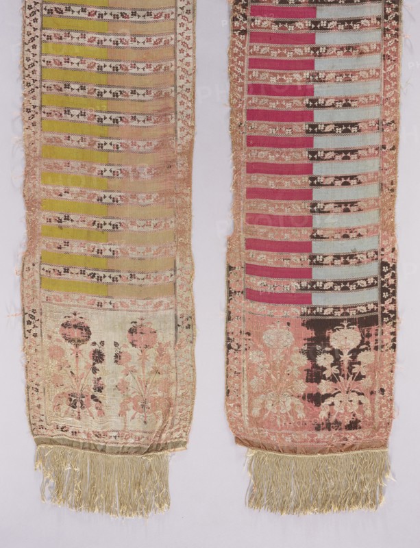 Silk kontusz sash, four-sided, c. 1787–1794. Likely made by Szczepan Filsjean Manufactory Kobylka. Bipartite ends each with a bouquet motif of rooted carnations tied with a ribbon. Field in transverse stripes alternating plain or two-coloured with fine floral tendrils; trim with small flowers. Borders vertical and horizontal with twig motifs and checkerboard. Sewn-on silk fringe. Colors: cream, brown, yellow-green, sky blue, dark red; right side: cream background of heads, borders and shelves with tendril, brown ornament; plain shelves half pink, half yellow-green; reverse: plain shelves half sky blue, half dark red. Weave density approx. 17/39 x2. Length 357 cm, width 32 cm. Origin: Kobylka, Masovian Voivodeship, Poland., Two long narrow textile panels hang side by side, each showing horizontal bands and floral motifs with fringed ends. The left panel features alternating horizontal stripes in shades of pale yellow and beige with narrow decorative bands in muted pink and dark brown, a...