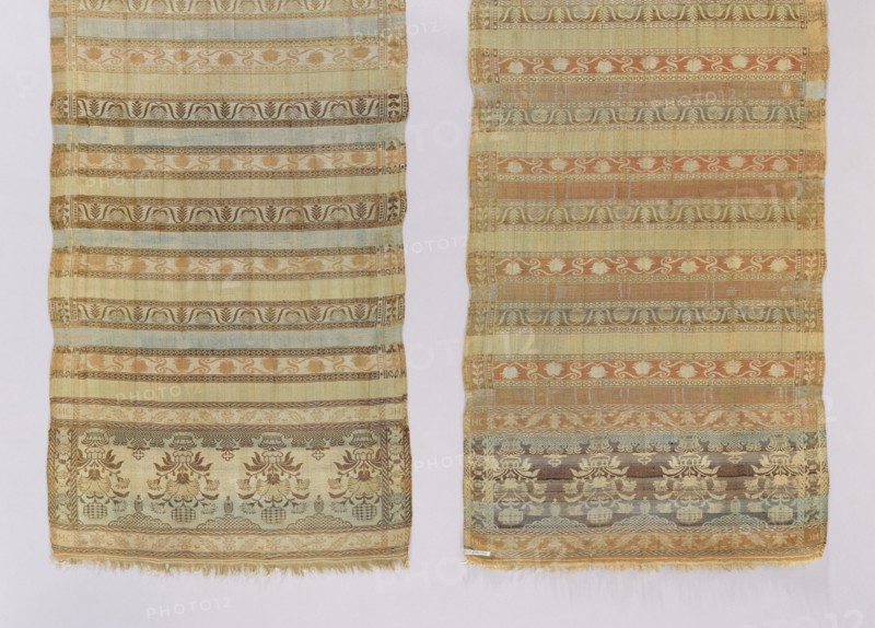 Kontusz belt. Quadripartite heads with a pattern of flower bouquets in inverted vases; earth marked at the bottom and clouds at the top. Alternating transverse stripes, smooth and varied in color, with tendril motifs in three varieties; partial edging with plant motifs. Colors: beige, brown, light blue, cream, pink, green. Reversible belt, men's clothing accessory. 2nd half of the 18th century, Volhynia. Length 290.5 cm; width 44.5 cm., Two long woven textiles are visible side by side against a pale background, each showing horizontal patterned bands with repeating motifs. The left textile has a muted palette of beige, light yellow, pale blue, soft brown, and faded green, with decorative floral and scroll-like designs and a fringe at the lower edge. The right textile features similar horizontal banding with colors including beige, warm tan, muted orange, pale blue, brown, and green, also showing floral and ornamental patterns and a slightly frayed lower edge. Both pieces display altern...