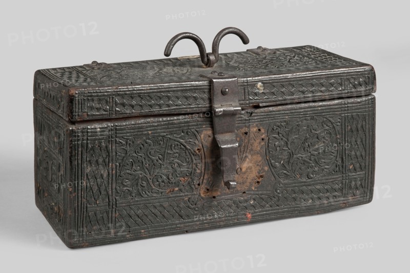 Casket, plant decoration, shields, dates, geometric ornament, early 16th century, possibly France, Flanders, or Germany; piece of furniture; height 12 cm, width 26.5 cm, depth 12.5 cm., A rectangular box with a hinged lid and a metal hasp and loop handle sits on a plain surface; the box is dark with worn areas revealing brown and reddish tones, and it is decorated across its surfaces with carved or embossed geometric and foliage patterns including braided borders and circular vegetal medallions, with a few small spots of lighter wear and rust around the metal fittings and hinge.