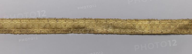 Five gallons, gallons haberdashery, origin unknown, geometric ornaments, 18th century, Europe, fabrics, width 1.5 cm, length 63 cm, A horizontal narrow woven band runs across the center of the image against a smooth background, showing a metallic golden color with subtle variations in tone; the band has a textured, patterned surface made of interlaced threads that create repeating oval and linear motifs, and its edges are slightly irregular with tiny loose fibers; the background is a uniform pale gray that contrasts with the warm gold of the band.