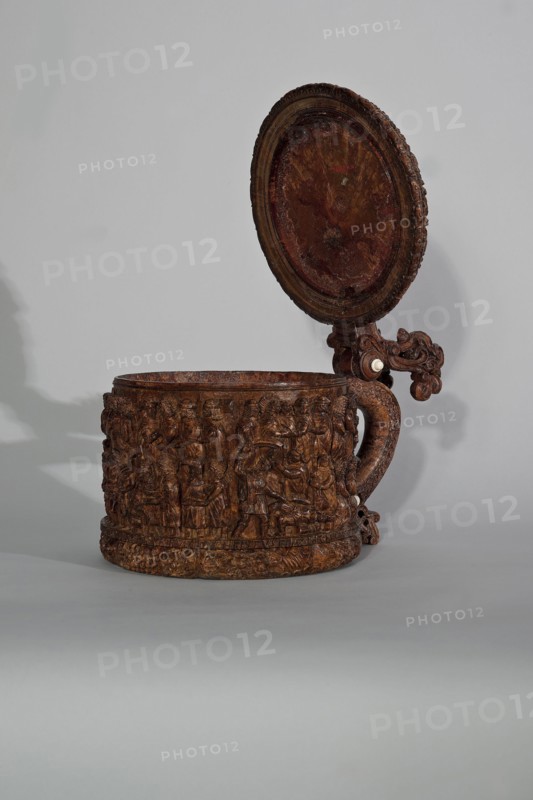 Wooden tankard, carved, probably a peg tankard, religious scenes, Gospel scenes, Resurrection of Christ iconography, inscriptions, vessels, Passion Bible; New Testament, human figures, figurative motifs, decorative art. Wooden pear-shaped tankard, so-called peg tankard, round, richly decorated with bas-reliefs depicting Passion scenes, with a Resurrection scene on the lid. The lid rim and handle are decorated with plant motifs and masks, and studded with ivory pins. In the center is a row of vertically driven metal pegs placed at regular intervals the top two are missing. 17th century, Scandinavia. Vessel. Height 23.5 cm, width 20.5 cm., A carved wooden tankard with an open lid sits against a plain light gray background; the tankard is a deep brown color with darker and lighter variations, and its cylindrical body is covered in intricate relief carvings depicting a group of human figures and decorative patterns, while the hinged circular lid shows worn textures and circular carving det...