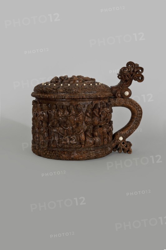 Wooden pear-shaped tankard, so-called peg tankard, carved, round, richly decorated with bas-reliefs depicting Passion scenes and a Resurrection of Christ scene on the lid; lid rim and handle decorated with plant motifs and masks and studded with ivory pins; center has a row of vertically driven metal pegs placed at regular intervals top two missing. Inscribed; figurative motifs of Gospel/Passion scenes and human figures. 17th century, Scandinavia. Vessel. Height 23.5 cm, width 20.5 cm., An intricately carved wooden vessel with a domed lid and a curved handle sits against a plain neutral background; the vessel displays detailed relief scenes around its cylindrical body and ornate scrolling decorations on the handle and lid, with small round insets visible on the lid and handle; the object is predominantly brown with lighter ivory-colored round accents and subtle variations in wood tone, set on a gray backdrop.