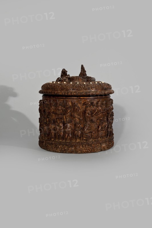 Wooden tankard, carved, probably a peg tankard, maker, religious scenes, Gospel scenes, Resurrection of Christ iconography; inscriptions; vessels; Passion Bible; New Testament; human figures; figurative motifs; ; decorative art. Wooden pear-shaped tankard, so-called peg tankard, round, richly decorated with bas-reliefs depicting Passion scenes, with a Resurrection scene on the lid. The lid rim and handle are decorated with plant motifs and masks, and studded with ivory pins. In the center is a row of vertically driven metal pegs placed at regular intervals the top two are missing. 17th century, Scandinavia; vessel. Height 23.5 cm, width 20.5 cm., A carved cylindrical container with a lid sits against a light gray background, the container rendered in rich brown tones with darker and lighter brown variations; the lid is topped by sculpted protrusions and a ring of small pale bead-like elements, and the body is covered in detailed low-relief carved figures and decorative motifs, casting ...