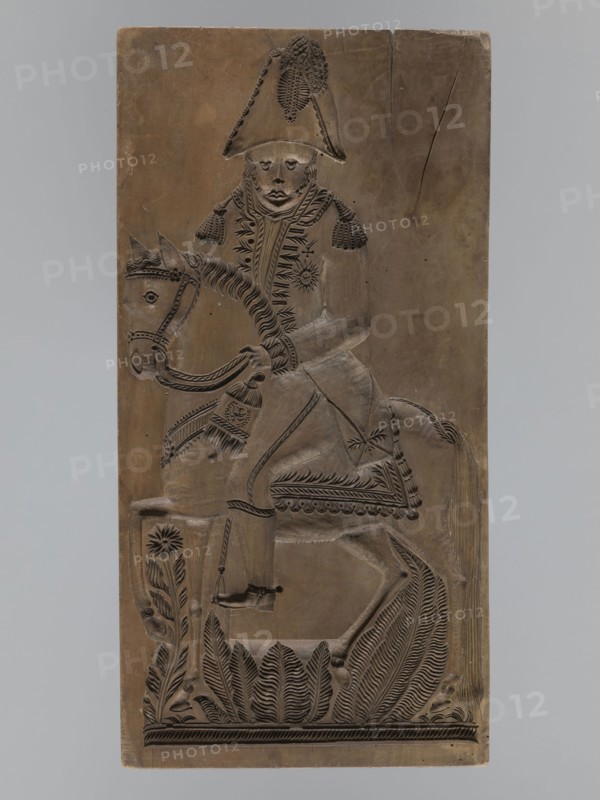 Gingerbread mold, double-sided. Obverse: image of a rider on a horse in a parade uniform from the Napoleonic era 1799–1815, wearing a bicorn, caftan with epaulettes decorated with braiding, an order on the chest, striped trousers and long boots; horse with decorative geometric motif; bottom decoration of leaves and flowers. Reverse: small image of a beehive. Rectangular wooden mold, edge carved with the letters CG Gl. initials. 18th–19th century, Province of Silesia 1742–1918. Height 51.5 cm, width 25.5 cm, depth 5 cm. Possible maker: woodcarver or gingerbread maker; subjects include Napoleonic soldiers, riders, floral decorations, historical military costume; decorative/applied art., A vertical rectangular carved panel showing a figure wearing a tall hat and ornate clothing seated on a horse; the carving is in shades of brown with darker incised lines forming details. The rider has a visible face with carved eyes, nose, and mouth and textured hair or headgear, and the coat features de...
