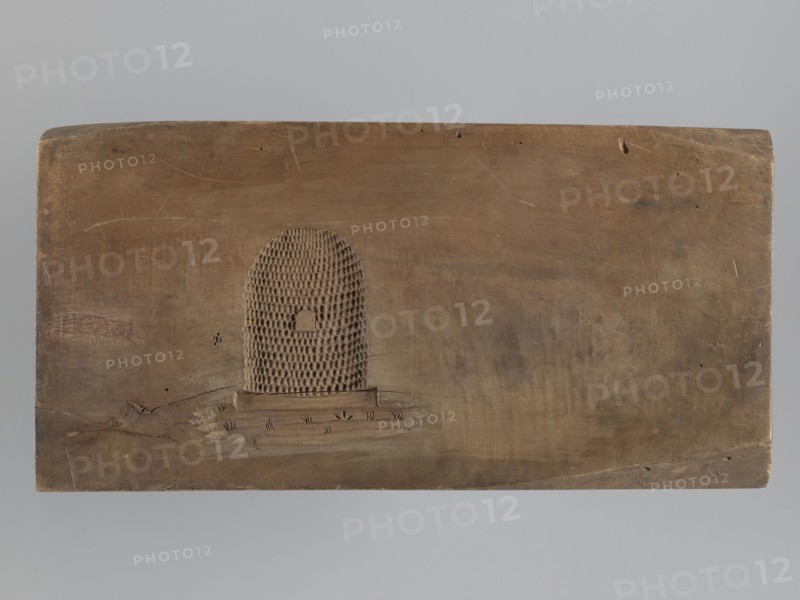 Gingerbread mold, double-sided. Obverse: image of a rider on a horse in a parade uniform from the Napoleonic Wars period, wearing a bicorn, caftan with epaulettes and braiding, an order on the chest, striped trousers and long boots; horse decorated with a geometric motif; bottom decoration of leaves and flowers. Reverse: small image of a beehive. Rectangular wooden mold, edge carved with the letters CG Gl. initials of the woodcarver or owner. Materials/technique: woodcarving for gingerbread molds; style: decorative/applied art; motifs: Napoleonic soldiers, riders, horse, floral decorations, historical military costume. Date: 18th–19th century. Place: Province of Silesia 1742–1918. Dimensions: height 51.5 cm, width 25.5 cm, depth 5 cm., A rectangular wooden board in shades of brown with a smooth, worn surface and visible wood grain, featuring a raised textured semicircular pattern near the center left composed of a honeycomb-like grid and a small arched cutout, a few shallow scratches a...