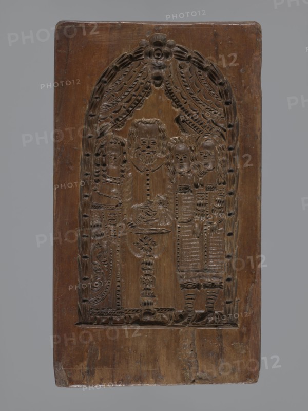 Double-sided gingerbread mould. Obverse: baptism scene; reverse: floral ornament. Genre scenes; gingerbread mould; folk-style decorations; baptisms; floral and plant motifs; applied decorative art; figurative scenes; historical costumes. Double-sided rectangular mould: on one side a genre scene depicting a baptism—group of figures in 17th–18th-century costumes; centre a bearded man, probably a priest or pastor, holding a child over a baptismal font, next to him three women in long patterned dresses; the whole group surrounded by an arched window decorated with rich geometric and plant ornament. On the other side a large leaf-shaped ornament containing interwoven floral and plant motifs. 18th–19th century, Province of Silesia 1742–1918. Form: height 26 cm, width 14.5 cm, depth 2.7 cm., A carved wooden panel with a rectangular shape and rounded top showing an arched carved scene framed by draped curtains and decorative motifs, carved figures standing under the arch including a central be...