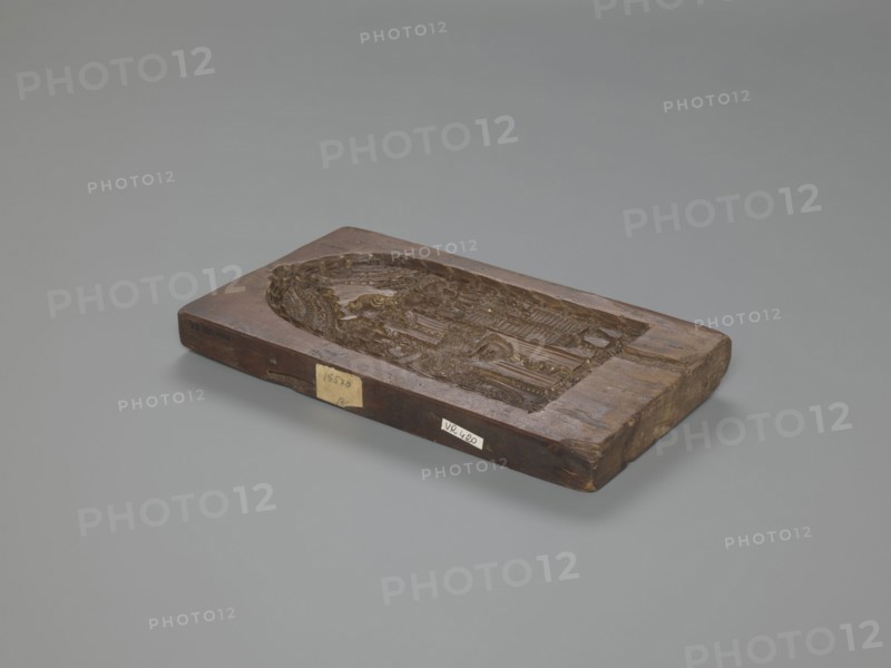 Double-sided gingerbread mould. Obverse: baptism scene; Reverse: floral ornament. Genre scenes, gingerbread moulds, folk-style decorations, baptisms, floral motifs, plant motifs, applied art, decorative art, gingerbread, figurative scenes, historical costumes. Double-sided rectangular mould: on one side a genre scene depicting a baptism — a group of figures in 17th–18th century costumes; in the centre a bearded man, probably a priest or pastor, holding a child over a baptismal font, next to him three women in long patterned dresses; the whole group surrounded by an arched window decorated with rich geometric and plant ornament. On the other side a large leaf-shaped ornament containing interwoven floral and plant motifs. 18th–19th century, Province of Silesia 1742–1918. Form: rectangular mould. Height 26 cm, width 14.5 cm, depth 2.7 cm., A rectangular wooden block with a carved recessed oval scene sits on a smooth grey background; the block shows a worn brown wood surface with darker an...