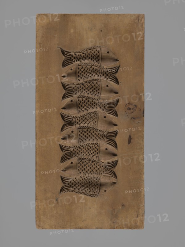 Double-sided gingerbread mould. Obverse: decorative pipe with fluted decoration; on the stem the letter A in an oval; at the mouthpiece two decorative tassels. Reverse: nine stylized fish covered with scales, arranged in a row. Folk-style decorations; applied/decorative art; gingerbread mould. 18th–19th century, Province of Silesia 1742–1918. Form rectangular; height 22.7 cm, width 11.5 cm, depth 3.5 cm., A rectangular wooden block with a vertical arrangement of a few carved fish motifs in relief running down its center, the carvings showing fish bodies with textured scale patterns and stylized fins and tails; the wood shows variations in tone and grain with knots and small imperfections, and colors visible include brown tones in the carved panel and a gray background surrounding the block.
