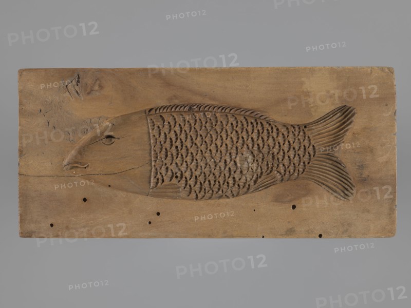 Double-sided gingerbread mold. Obverse: baby in swaddling clothes richly decorated with various geometric motifs; reverse: stylized elongated fish covered with scales. Gingerbread mold; folk-style decorations; applied/decorative art; folk customs; geometric ornaments; children; fish. 18th–19th century, Province of Silesia 1742–1918. Material: mold for gingerbread. Dimensions: height 16.7 cm; width 7.8 cm; depth 3.4 cm., A rectangular wooden panel with a carved fish motif occupies the center, the fish rendered in shallow relief with a rounded head, a carved eye and mouth, a textured body of repeating scale-shaped cutouts, and a fan-shaped tail with carved striations; the panel shows natural wood grain, knots, small holes, and areas of darker and lighter brown, set against a smooth gray background.