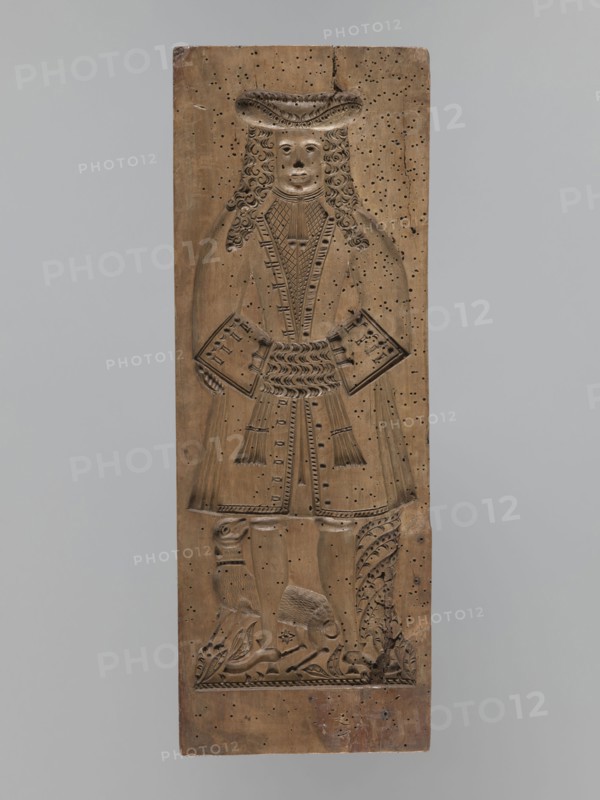 One-sided gingerbread mould with the image of a bachelor;, Rectangular mould showing a male figure standing with joined hands, wearing a patterned tricorn hat and a long wig, in a bachelor’s costume from the 2nd half of the 19th century. Attire includes a justokor, culottes, stockings and high-heeled shoes. A dog stands at the man's leg. Decorative plant motifs at the bottom. Folk-style decorations; applied and decorative art. Materials: gingerbread mould. Date: 17th century or early 18th century; also associated with 18th–19th century. Region: Silesia Province of Silesia, 1742–1918. Dimensions: height 50 cm, width 18 cm, depth 4.2 cm., A tall vertical wooden panel carved in a warm brown tone shows a full-length figure of a person in period dress with long curly hair and a wide brimmed hat, hands held at the waist holding rectangular objects, and a long coat with decorative carved patterns; the lower portion of the panel features carved legs and shoes above a carved ground with floral ...