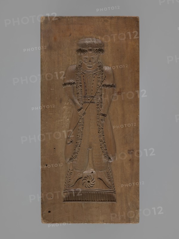 Double-sided gingerbread mould. Obverse: female figure in a long, patterned, richly decorated dress, wearing a hat decorated with an egretta. Reverse: male figure with a fur hat, dressed in a fur-lined coat over a shirt with decorative embroidery, holding a long pipe. Plant decoration at the bottom of both figures. Woodcarving; folk-style decorations; applied/decorative art; female and male figures; historical costumes; water pipes. 18th–19th century, Province of Silesia 1742–1918. Double-sided rectangular mould. Height 19 cm, width 9 cm, depth 3.3 cm., A carved wooden panel showing a full-length figure in relief standing on a decorated rectangular base; the figure has long curled hair or braided detailing, a round hat or headpiece, and wears a long garment with decorative edging and textured patterns; the figure’s arms are bent at the elbows with hands held near the waist, and a thin carved element extends diagonally across the torso; the panel surface shows natural wood grain, small ...