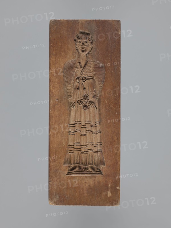 Double-sided gingerbread mould. Obverse: female figure in a long, patterned, richly decorated dress, wearing a hat decorated with an egretta. Reverse: male figure wearing a fur hat and a fur-lined coat over an embroidered shirt, holding a long pipe. Plant decoration at the bottom of both figures. Woodcarving, folk-style decorations; gingerbread moulds; decorative/applied art; historical costumes. 18th–19th century, Province of Silesia 1742–1918. Rectangular form; height 19 cm, width 9 cm, depth 3.3 cm., A rectangular wooden block carved in relief with the figure of a standing person wearing a long dress and a headpiece; the wood shows a warm brown tone with lighter and darker variations and areas of wear, and the background around the carving is a similar brown; the carved figure has textured details on the sleeves and skirt, a belt with a tied bow, and floral-like shapes held near the waist, with carved folds at the hem and a small raised base beneath the feet; the overall scene is se...