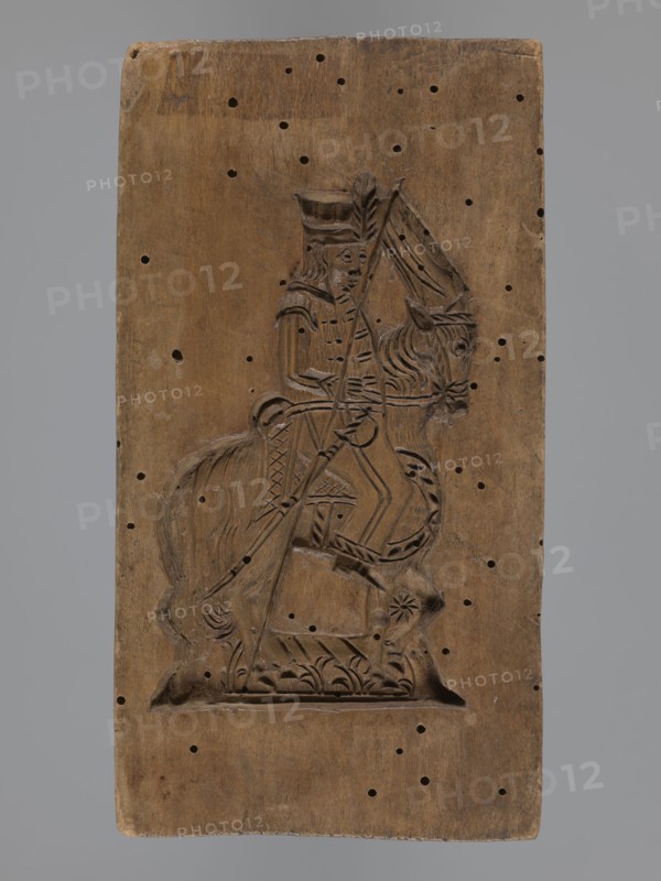 Double-sided gingerbread mould with the image of a rider on both sides, gingerbread; subject: horses, riders in historical military costumes hussar-type and another military uniform, sabres, pole weapon lance, horse head with decorative covering; style: folk-style decorations, plant motifs; materials/function: gingerbread mould, decorative art; form: double-sided, rectangular mould; inscriptions/details: one side—rider in military uniform decorated with braiding, high mirliton-type headgear, holding a sabre, plant decoration at the bottom; other side—rider in a different military uniform wearing a cap with a feather, holding a pole weapon, sabre at his side, horse head decorated, decorative plant motifs at the bottom; date: 18th–19th century; place: Province of Silesia 1742–1918; dimensions: height 17.2 cm, width 9 cm, depth 2 cm., A rectangular wooden panel with a carved relief of a mounted figure occupies the center; the wood is brown with a weathered surface and shows a pattern of s...
