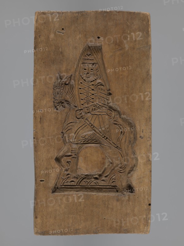 Double-sided gingerbread mould with the image of a rider on both sides;, Gingerbread; folk-style and plant decorations; horses, uniforms, sabres, plant motifs; hussars; horse rider iconography; historical costumes. Rectangular mould: one side shows a rider in a military uniform decorated with braiding, likely a hussar, wearing a high mirliton-type headdress and holding a sabre, with plant decoration at the bottom. The other side depicts a rider in a different military uniform, wearing a cap with a feather, holding a pole weapon lance, with a sabre at his side and the horse’s head covered decoratively; decorative plant motifs at the bottom. 18th–19th century, Province of Silesia 1742–1918. Form: double-sided. Height 17.2 cm; width 9 cm; depth 2 cm., A rectangular wooden panel with a carved relief of a mounted figure occupies the center. The panel is brown with lighter and darker wood grain variations. The carved scene shows a person seated on a horse; the person wears a pointed headpiec...