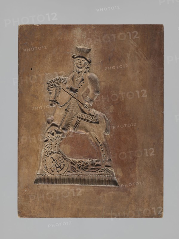 Double-sided gingerbread mould. Rectangular wooden form; obverse: image of a lion with abundant mane, standing on a base decorated with plant-twig ornament, holding a ball engraved with the date 1829 and a symbol of an anchor intertwined with the letter S; bottom with floral and plant decoration. Reverse: figure of a cavalier on horseback in early 19th-century riding attire jacket, breeches, top hat, holding a riding crop; at the horse's front legs a round shield engraved 1831 and the same anchor-with-S symbol mark of the woodcarver or owner; bottom with floral and plant ornament. Materials: wood gingerbread mould; subject matter: rider on a horse, lion, figurative scenes, plant motifs; style: applied folk/culinary mould; date marks: 1829 and 1831; region: Silesia Province of Silesia, 1742–1918. Dimensions: height 25.8 cm, width 19.4 cm, depth 4.3 cm., A rectangular wooden panel with a carved relief of a mounted figure occupies the center; the panel shows a warm brown wood tone with li...