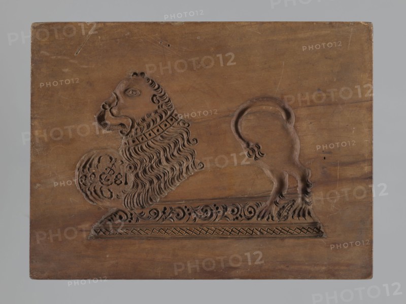 Double-sided gingerbread mould. Rectangular form, height 25.8 cm, width 19.4 cm, depth 4.3 cm. Obverse: image of a rider — figure of a cavalier on horseback in early 19th-century riding attire jacket, breeches, top hat, holding a riding crop; at the horse’s front legs a round shield engraved “1831” and a mark showing an anchor intertwined with the letter “S”; bottom decorated with floral and plant ornament. Reverse: stylised lion with abundant mane standing on a base decorated with plant-twig ornament, holding a ball engraved “1829” and the same anchor-with-S mark; bottom with floral and plant decoration. Materials/technique: carved mould for gingerbread applied art. Iconography: rider on a horse, lions, plant motifs; historical costumes. Place/date attribution: Silesia, Province of Silesia 1742–1918; dates on images 1829 and 1831., A carved wooden panel shows a stylized animal with a large mane and a curled tail, rendered in relief against a plain wooden background; the carving includ...