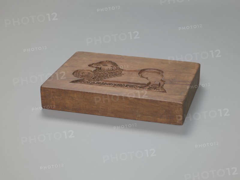 Double-sided gingerbread mould. Rectangular shape, height 25.8 cm, width 19.4 cm, depth 4.3 cm. Obverse: image of a rider on horseback in early 19th-century riding attire jacket, breeches, top hat, holding a riding crop; at the horse's front legs a round shield engraved 1831 and an anchor intertwined with the letter S mark of the woodcarver or owner; bottom floral and plant ornament. Reverse: stylised lion with abundant mane standing on a base decorated with plant twigs, holding a ball engraved 1829 and the same anchor entwined with S; bottom floral and plant decoration. Materials/function: carved gingerbread mould; motifs include figurative scenes, lions, horse, historical costumes, and plant motifs. Region/date: Silesia, Province of Silesia 1742–1918; dates on mould: 1829 and 1831., A rectangular wooden block in brown tones rests on a smooth pale gray surface; the top face of the block features a carved raised design of an animal-like figure with a mane and curled tail plus decorativ...