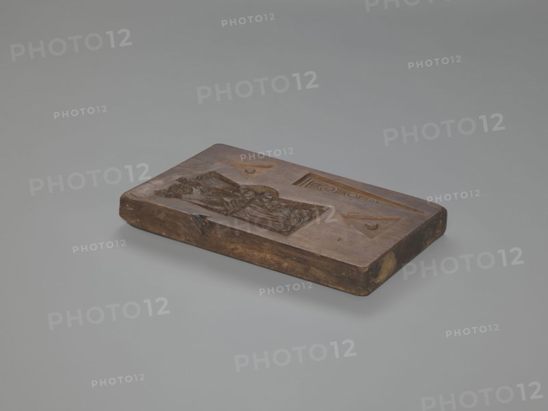 Double-sided gingerbread mould. Obverse: Easter Lamb holding a banner, haloed, with a chalice collecting flowing blood, background strewn with numerous flowers, surrounded by a wreath woven from laurel leaves. Reverse: female figure seated at a spinning wheel in a long decorative dress with a deep neckline, plus three triangular motifs decorated with geometric elements. Rectangular mould, four motifs on one side. Material: wood woodcarving; function: gingerbread mould. Style/subjects: religious motifs Lamb of God, festive traditions, spinning wheel, female figure, laurel wreath, floral and geometric ornament. Date: 18th–19th century. Region: Province of Silesia 1742–1918. Dimensions: height 28 cm, width 14.4 cm, depth 2.5 cm., A rectangular carved wooden block rests on a smooth pale gray surface against a lighter gray background; the block is a warm brown with darker brown areas and some lighter tan spots at the edges, and its top surface features recessed carved shapes including a rec...