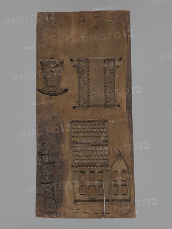 Double-sided gingerbread mould. Obverse: figure of a bearded man in patterned clothes, richly decorated with geometric motifs, wearing a hat with a feather and blowing a horn French horn; plant ornament at the bottom. Reverse: several small elements including houses with tiled roofs, a cradle decorated with a flower motif, and a chest or bed decorated with a plant twig. Folk-style decorations, figurative scenes; geometric and plant ornaments; male figure; applied/decorative art. Double-sided rectangular form. 18th–19th century, Silesia Province, 1742–1918. Height 30 cm, width 13.8 cm, depth 2 cm., A vertical wooden panel with carved depressions showing a variety of decorative motifs and architectural forms, the wood ranging from medium brown to darker brown patches and a strip of rough, cracked dark brown bark along one side; near the top a carved band with ornate vertical panels and scrolling leaf patterns sits beside a shallow curved floral motif, while below there is a carved rectan...