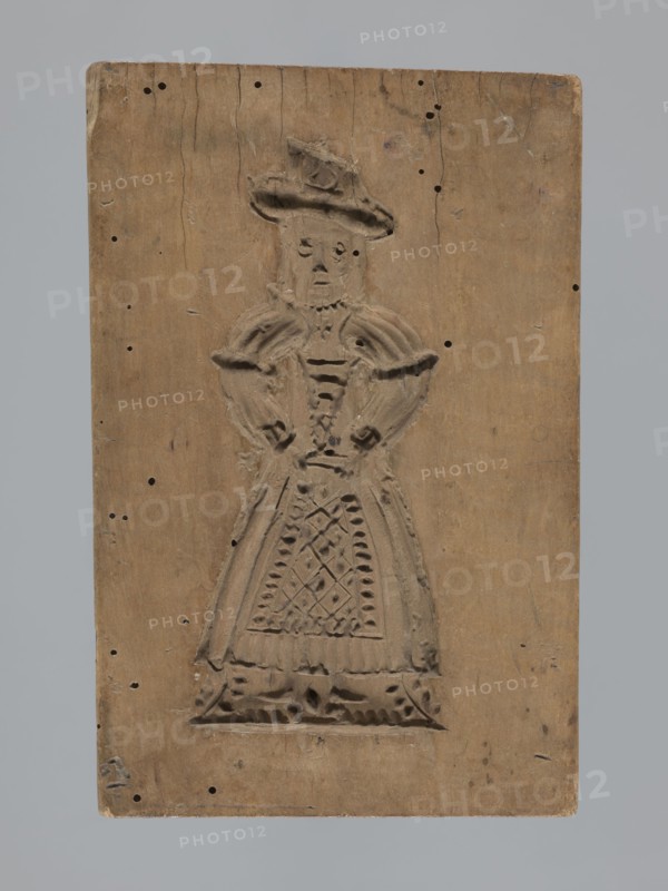 Double-sided gingerbread form. Obverse: female figure in a long patterned dress wearing a hat. Reverse: four images — a baby in swaddling clothes, a fish, and two shoes. Rectangular form, applied/folk decorative art with figurative and zoomorphic motifs; female figures in rich dresses; folk customs. Gingerbread mold, 18th–19th century, Province of Silesia 1742–1918. Height 17 cm, width 11 cm, depth 2 cm., A rectangular wooden board in brown with a carved relief of a figure wearing a hat and a long dress occupies the center; the carved figure shows textured details on the hat, shoulders, bodice, and a patterned apron with lattice-like carving. The wood surface shows a few small dark holes and tiny darker blemishes scattered across it, along with hairline cracks and surface wear; the board edges are slightly rounded and worn. The background around the board is a uniform light gray. The visible colors are brown on the wooden board and light gray in the surrounding background.