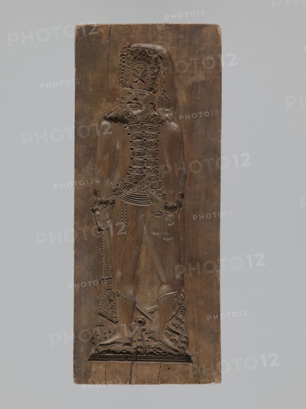 Double-sided gingerbread form. Obverse: male figure with a moustache in a hussar uniform, wearing a fur-lined jacket, standing with legs slightly apart, leaning on a sabre held in his right hand, wearing a high shako topped by a two-headed eagle and tassel; plant ornament at the bottom. Reverse: stylized elongated fish covered with scales, depicted in profile. Rectangular form, applied art/gingerbread mold, decorative folk motifs, soldiers/hussars, historical costumes. 18th–19th century, Province of Silesia 1742–1918. Height 28.7 cm; width 11.7 cm; depth 3.5 cm., A carved wooden panel in brown tones shows a relief of a standing human figure with detailed textured hair and a tall hat, wearing a textured upper garment and a belt, with visible sculpted hands and feet, and holding a long object that reaches to the base; the panel background is smooth with variations in brown, and the image also shows a pale gray background surrounding the panel.