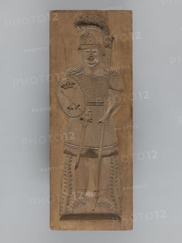 Double-sided gingerbread form; obverse: female figure in a helmet; reverse: male figure in a helmet. Gingerbread form, applied/decorative art; materials: gingerbread. Description: Double-sided rectangular form — one side shows a female figure in a long patterned dress with a neckline, wearing a helmet decorated with a plume, holding a sword in one hand and a roll of paper in the other, wearing shoes, with a plant ornament at the bottom. The other side shows a male figure in armor resembling Roman legionary or Greek hoplite armor, wearing a similar helmet with plume, holding a shield bearing two crossed swords and a crown and a javelin in the other hand, a sword girded at his side, wearing sandals, with a plant ornament at the bottom. Themes: militaria, historical costumes, armour, swords, male and female figures, plant ornaments, decorative/applied art. Date: 18th–19th century. Place: Silesia Province 1742–1918. Type: form. Dimensions: height 59 cm; width 22 cm; depth 4.5 cm. Creator: ...