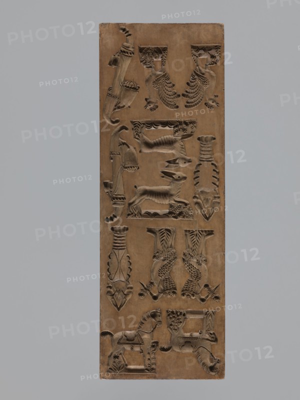 Double-sided gingerbread mould, maker signed Glauer woodcarver or gingerbread maker, dated j.1827. Rectangle mould, height 29 cm, width 10.5 cm, depth 2.4 cm. One side: twelve small images of animals — two horses, two roosters, two crayfish, and two storks carrying babies in baskets — and two decorative pipes. Other side: miniature elements including children in swaddling blankets, shoes, footwraps, a cradle, a guitar, and additional motifs adorned with plant and geometric ornaments. Materials/genre: carved wooden form for gingerbread; folk motifs; zoomorphic and anthropomorphic figures; geometric and plant ornamentation. Silesia Province of Silesia, 1742–1918; dated 1826–1827., A vertical rectangular wooden panel carved with a repeating pattern of decorative relief motifs including stylized animals, birds, a horse, and abstract ornamental shapes, all in shades of brown; the background around the panel is a solid light gray.