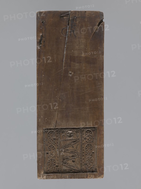 Double-sided gingerbread form. Obverse: figure of an angel with the alphabet. Reverse: various floral ornaments. Double-sided rectangular form; on one side a male figure probably an angel standing, in a costume richly decorated with plant ornament, girded with two sashes, holding a decorative board with the alphabet inscribed; above the figure's head a halo; in the background a feather and flame motif possible angel wings. The other side: rectangular field filled with floral and plant decoration, including the head of a baby. At the top a signature: initials G and R mark of the woodcarver or gingerbread maker/workshop owner. 18th–19th century, Province of Silesia 1742–1918. Material/type: form. Height 32.3 cm, width 13 cm, depth 2 cm., A tall rectangular wooden panel with a dark brown surface showing a vertical crack and several small dents and scratches, with a carved rectangular decorative panel near the bottom featuring floral and foliate motifs and a small carved face, set against ...