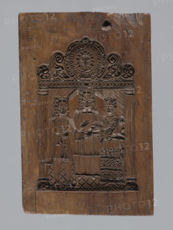 A single-sided gingerbread mould with a wedding ceremony scene, figurative scenes, gingerbread mould, weddings, male figures, female figures, volutes, folk-style decorations, plant motifs, gingerbread, Silesia Province of Silesia; 1742–1918, decorative art, folk motifs. A single-sided rectangular mould with a wedding scene: in the centre a priest dressed in a chasuble, wearing a wig; to his left a man and to his right a woman with their hands joined by the priest. Both wear richly decorated turn-of-the-17th/18th-century costumes and wigs — the man in a justaucorps, culottes and stockings, the woman in a long patterned dress with a low neckline. Above the heads of the figures is a decorative finial supported by two columns; its central element is a round clock face showing a sun with a human face surrounded by volute and plant ornament, hands on the face, and a rim marked with Roman numerals. Date: 18th–19th century. Dimensions: height 23 cm, width 15.5 cm, depth 4.5 cm., A rectangular ...